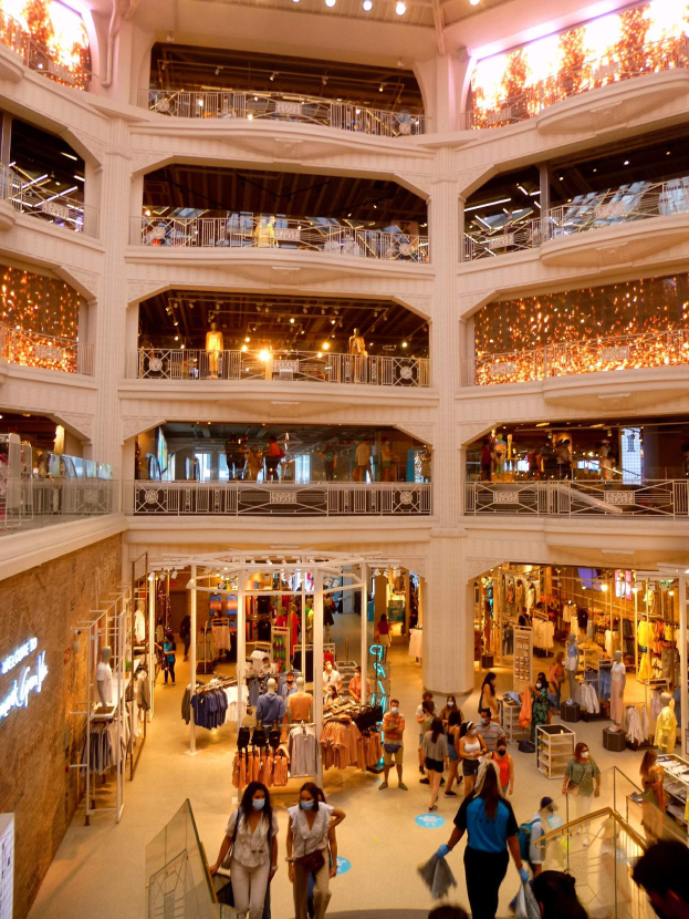 Ein belebter Einkaufszentrumsvorplatz mit Menschen, Schaufensterpuppen mit Kleidern, Geländern, Lampen und Wandtafeln.