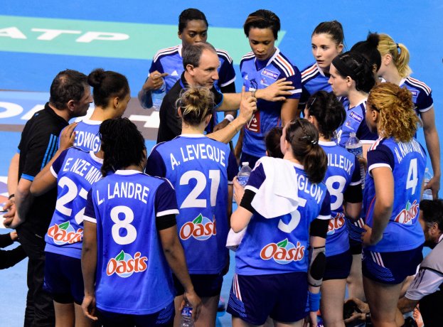 Eine Gruppe von Frauen-Handballspielerinnen steht zusammen auf einem Feld, einige halten Flaschen, während jemand mit einer Kamera das Geschehen auf der rechten Seite festhält.