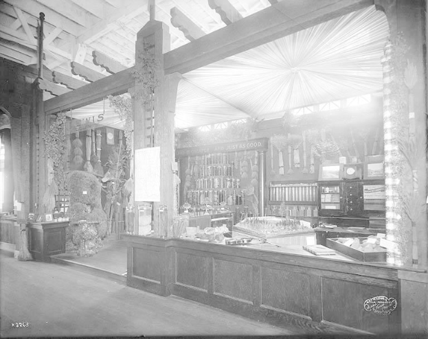 Ein altes Schwarz-Weiß-Foto einer Apotheke mit einem Tresen, der verschiedene Gegenstände zeigt, Regalen mit Flaschen und anderen Gegenständen im Hintergrund.