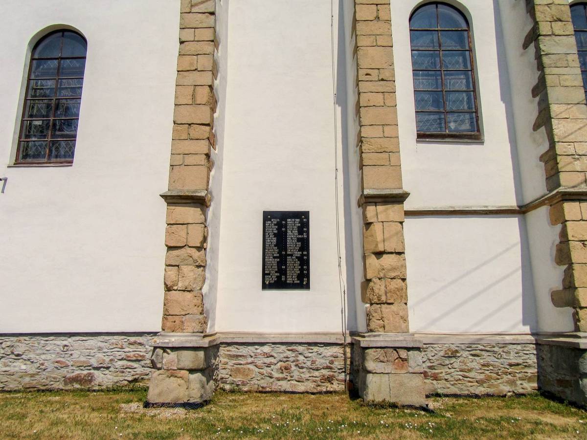 Eine weiße Kirche mit einer Steinmauer und Fenstern, umgeben von Gras und einem klaren blauen Himmel, zeigt eine Gedenktafel für die Opfer des Holocaust.