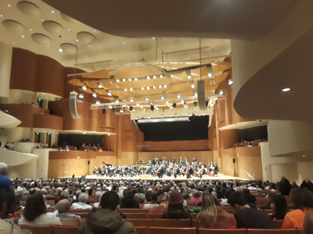 Eine große Halle voller Menschen, die ein Konzert anschauen, mit einer Gruppe auf der Bühne, die Instrumente spielt und anderen, die sitzen, unter einer Decke, die mit Lichtern geschmückt ist.