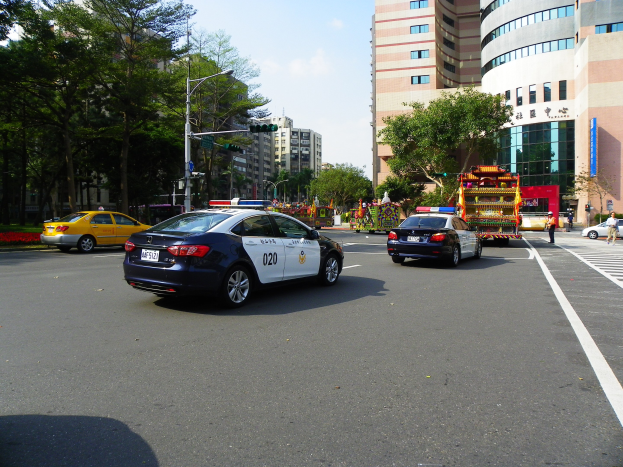 Ein Polizeiwagen fährt eine Straße mit hohen Gebäuden, Bäumen, Laternen, Verkehrsampeln und Menschen unter einem bewölkten Himmel entlang.