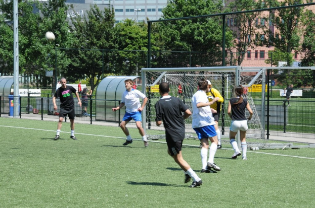 Menschen spielen Fußball auf einem Feld mit einem Zaun und einem Tor in der Mitte, einem Pfahl und einem Ball oben links und Bäumen und Gebäuden im Hintergrund.