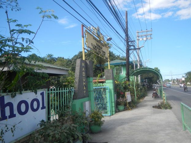 Der Eingang einer Schule mit einem Schild, auf dem "Schule" steht, auf der Seite einer Straße, mit einem Torbogen, Tor, Namenstafel, Pflanzen, Bäumen, Strommasten mit Drähten und einem bewölkten Himmel im Hintergrund.