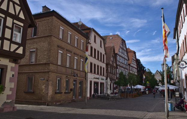 Eine Kopfsteinpflasterstra├če in Rothenburg ob der Tauber, Deutschland, gesäumt von Geb├Ąuden, B├Ąumen und Fahnen, mit Menschen und Fahrzeugen auf der Straße unter einem bew├Âlktem Himmel.
