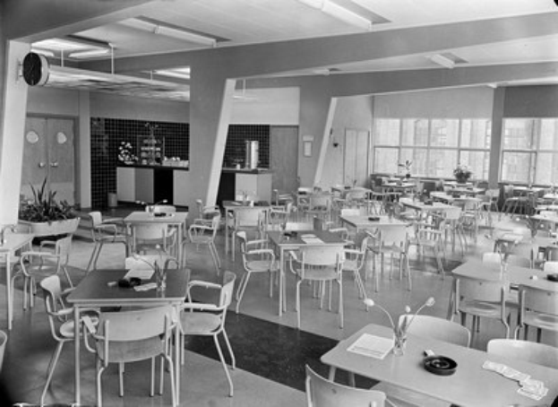 Schwarz-weißes Foto einer Cafeteria mit Tischen und Stühlen, Tellern, Besteck, Gläsern, Blumenvasen und Deckenleuchten; Türen und Fenster säumen die Hintergrundwand.