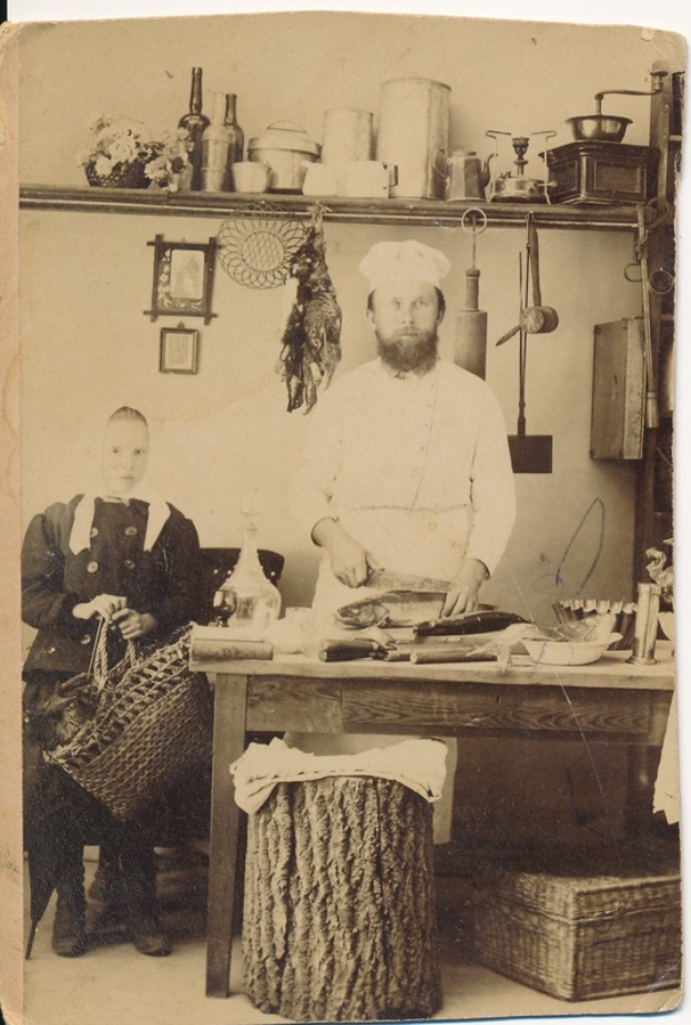 Ein altes Foto eines Mannes in einer Küche mit einem Messer in der Hand und einer Frau auf einem Stuhl mit einem Korb, umgeben von verschiedenen Gegenständen auf dem Tisch und Rahmen an der Wand.