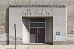 Das Südliche Berufsbildungszentrum mit einer Tür, Schildern an der Wand und einem Weg, der zur Eingangstür führt.