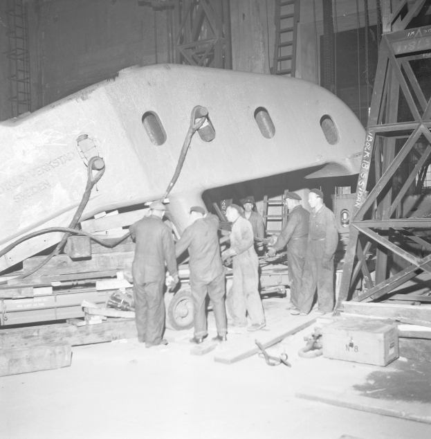 Eine Gruppe von Männern in Mützen arbeitet an einem Flugzeug in einer schwarzen und weißen Fabrikumgebung, umgeben von Metallobjekten, Leitern und anderen Gegenständen.