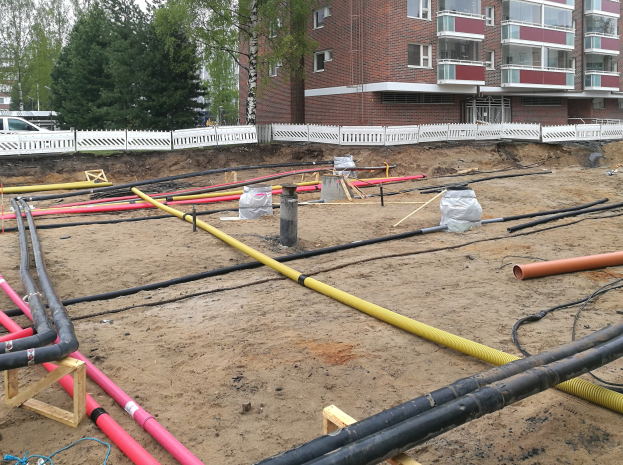 Großer Haufen von Rohren auf einer Baustelle mit verstreuten Materialien, umgeben von Gebäuden und Bäumen unter einem klaren Himmel.