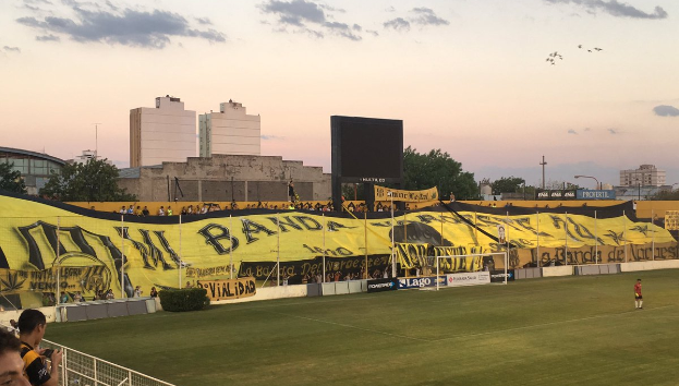 Ein Fußballfeld mit einer Gruppe von Menschen darauf, umgeben von einem Metallzaun, einem Torpfosten, einer Fahne mit der Aufschrift "Boca Juniors vs Real Sociedad", einigen Pflanzen, einem Anzeigebildschirm, einigen Pfosten, der Rinde eines Baums, einigen Bäumen, einigen Gebäuden und einem bewölkten Himmel.