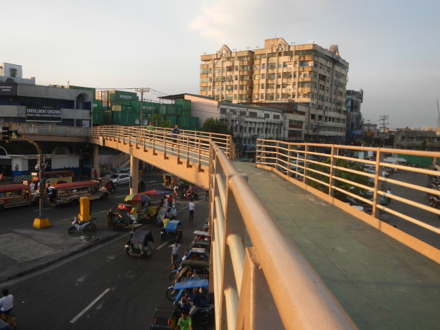 Eine Brücke überspannt eine belebte Straße mit darunter fahrenden Fahrzeugen, Fußgängern und Schirmen, flankiert von Geländern und vor dem Hintergrund von Gebäuden, Bäumen, Pfählen und einem klaren blauen Himmel.