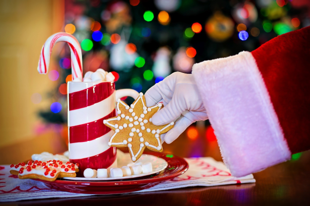 Eine Person mit einem Santa Claus Hut, die einen Becher heiße Schokolade und einen Zuckerstange hält und ein Teller mit Keksen und ein Tuch auf dem Tisch vor sich hat, vor einem Weihnachtsbaum und einer Wand.