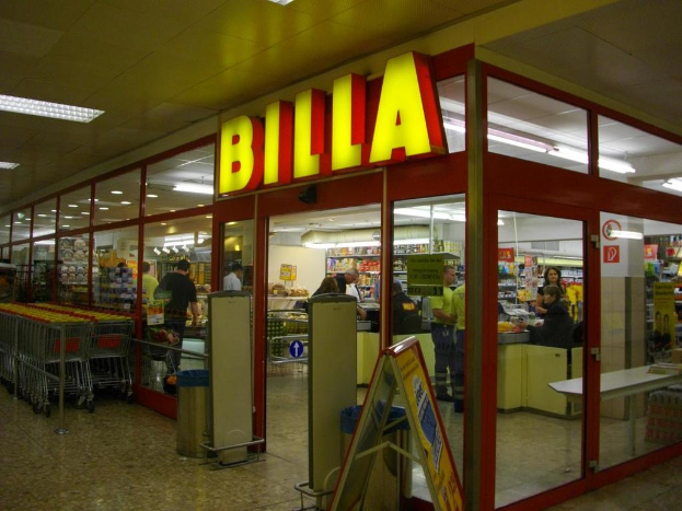 Ein Markt mit einem Eingang in der Mitte, Mülltonnen daneben, eine Reihe von Einkaufswagen auf der linken Seite, Deckenbeleuchtung und einige Menschen, die Produkte durchstöbern.
