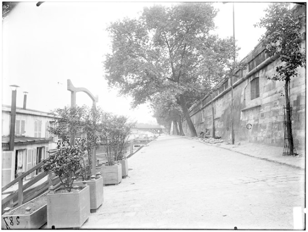 Eine schwarz-weiße Straßenansicht des New Yorker Staatsgefängnisses mit einer Wand auf der rechten Seite, Bäumen und Pflanzen auf der linken Seite und einer Brücke im Hintergrund.