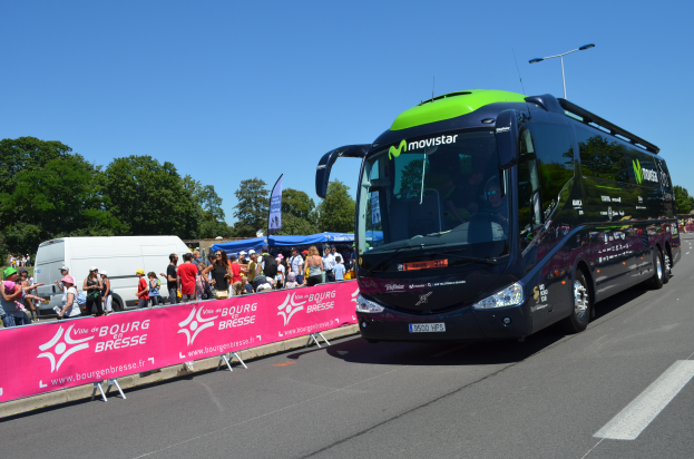 Ein schwarzer und grüner Bus fährt auf einer Straße neben einer Menge Menschen, einige tragen Mönen, mit einem Banner auf der linken Seite und Bäumen unter einem klaren blauen Himmel im Hintergrund.