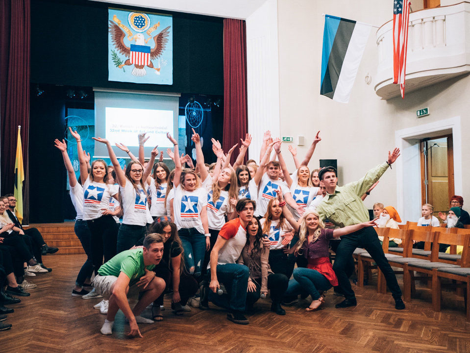 Eine Gruppe von Menschen, die vor einem Publikum posiert, das auf Stühlen sitzt und einige stehen, mit einem Bildschirm, Vorhängen, Flaggen, einem Schild, einer Tür, einem Geländer und einer Decke mit Deckenleuchten im Hintergrund.