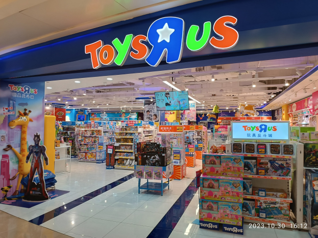 Ein Toys R Us Geschäft in einem Einkaufszentrum mit Regalen voller Spielzeug, einem Namensschild mit Text, Deckenlampen und einem Wasserzeichen in der rechten unteren Ecke.