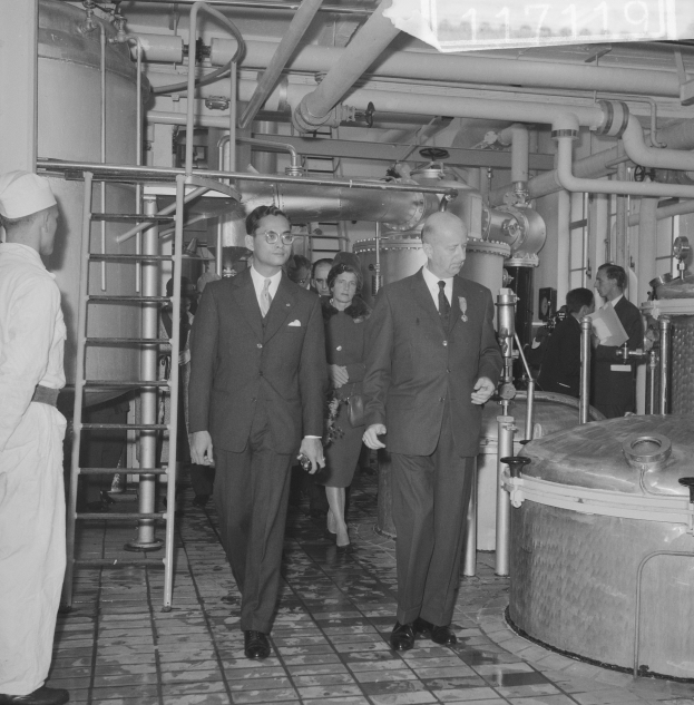 Eine Gruppe von Männern in Anzügen und Krawatten geht durch eine schwarz-weiße Fabrik, mit einer Leiter links und Maschinen im Hintergrund.