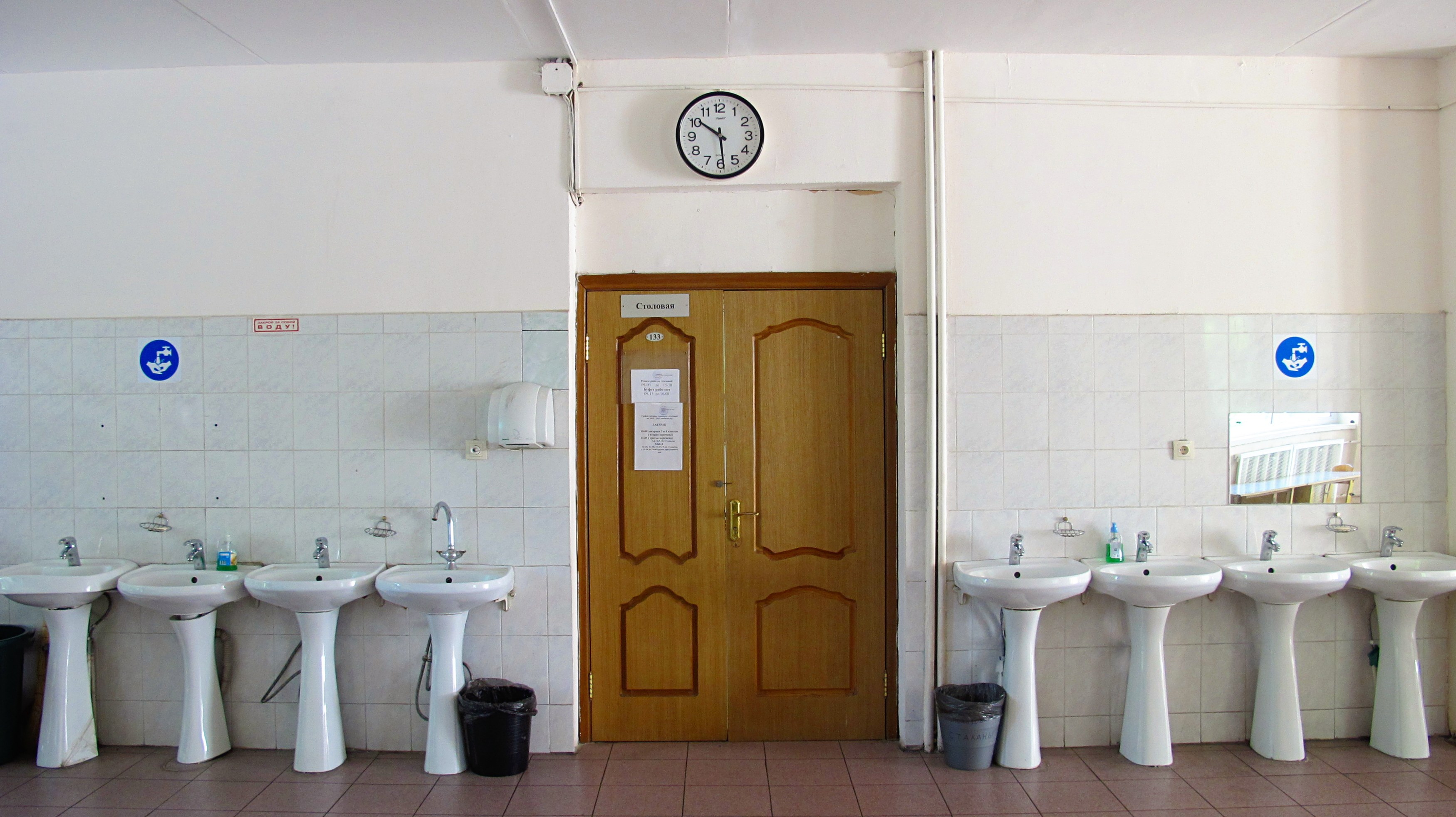 Ein Badezimmer mit einer Reihe von Waschbecken mit Wasserhähnen, Mülltonnen auf dem Boden, einer Tür im Hintergrund, einer Uhr an der Wand und einem Spiegel auf der rechten Seite.