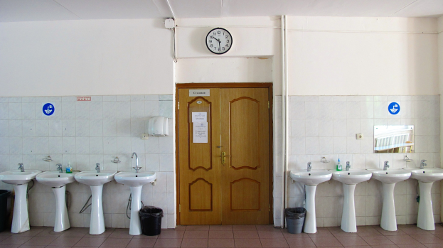 Ein Badezimmer mit einer Reihe von Waschbecken mit Wasserhähnen, Mülltonnen auf dem Boden, einer Tür im Hintergrund, einer Uhr an der Wand und einem Spiegel auf der rechten Seite.