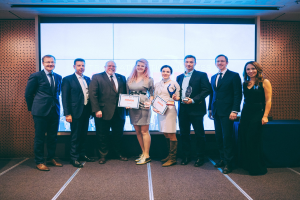 Gruppe von strahlenden Menschen mit Urkunden vor einem Bildschirm, mit einem Tisch und einem wandmontierten Objekt im Hintergrund, Gewinner der International Business Awards 2018.