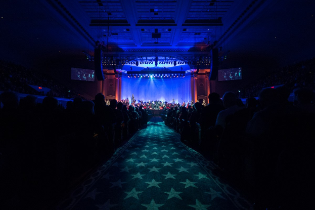 Ein großer Saal mit Stühlen, auf denen Menschen sitzen, die einer Bühne mit Musikern zugewandt sind, mit Deckenleuchten und Fernsehern, was auf ein Konzerthaus während der Nationalkonvention 2019 hinweist.