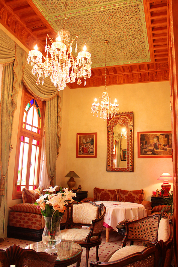Ein Wohnzimmer mit einem Tisch und Blumentopf im Vordergrund, umgeben von Stühlen, Sofas und einem mit einer wei√üen Decke bedeckten Tisch im Hintergrund, gerahmte Bilder an der Wand, von der Decke hängende Lampen und Vorhänge mit einem Fenster links.