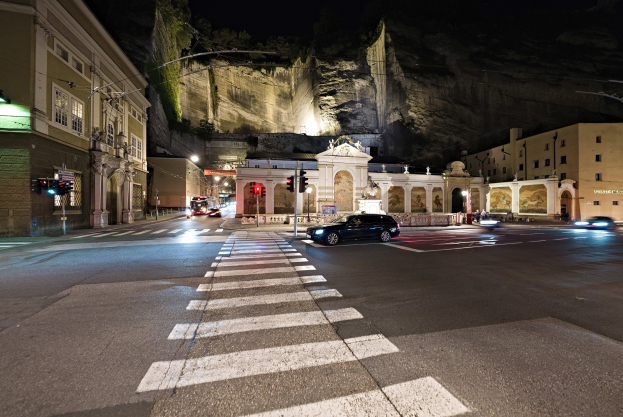 Eine nächtliche Stadtstraße mit fahrenden Autos, Straßenlaternen, Ampeln, beleuchteten Gebäuden, Laternenpfählen, einem Hügel im Hintergrund und einer Zebrastreifenmarkierung am Boden.