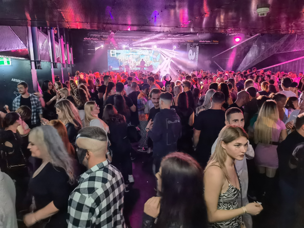 Eine große Gruppe von Menschen in einer Diskothek, beleuchtet von Deckenlichtern und umgeben von Säulen, mit einem Bildschirm im Hintergrund, der ein Absolut Nightlife-Event suggeriert.