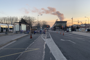 Eine Stadtstraße mit Verkehrskegeln, einem Bus, Laternenpfählen, Bäumen, Gebäuden, Kränen und Rauch, der aus einem fernen Gebäude aufsteigt.