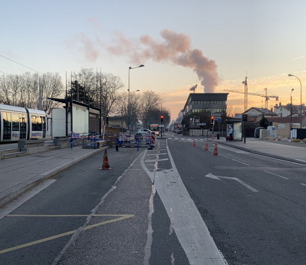 Eine Stadtstraße mit Verkehrskegeln, einem Bus, Laternenpfählen, Bäumen, Gebäuden, Kränen und Rauch, der aus einem fernen Gebäude aufsteigt.