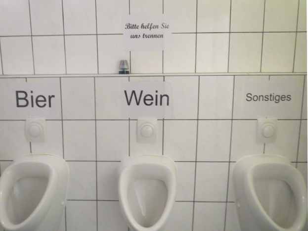 Drei weiße Toiletten an einer Wand montiert, jeweils mit der Aufschrift "Bier Wein" oder "Sonstiges".