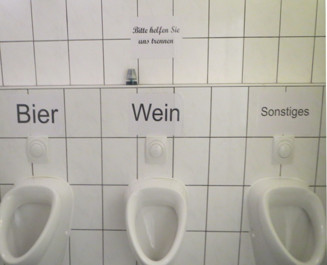 Drei weiße Toiletten an einer Wand montiert, jeweils mit der Aufschrift "Bier Wein" oder "Sonstiges".
