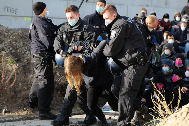 Gruppe maskierter Personen in Mützen, einige bewaffnet, die eine Frau umgeben, die von der Polizei eskortiert wird, mit Pflanzen, Steinen und protestrelevanten Wandtexten im Hintergrund.