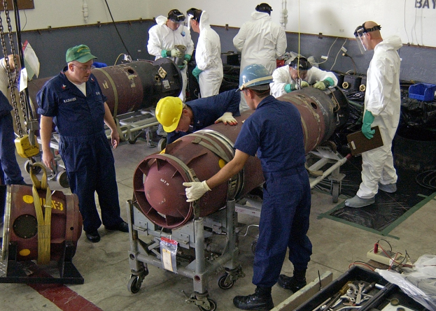 Eine Gruppe von Männern in Helmen und Handschuhen demontiert eine große Bombe in einer Fabrik mit Maschinen und Text an der Wand.