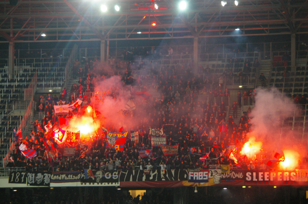 Eine große Menschenmenge in einem Stadion hält Fahnen und Banner, mit Leuchtraketen, die Rauch erzeugen, unter einer Decke mit Lampen und Metallrahmen.
