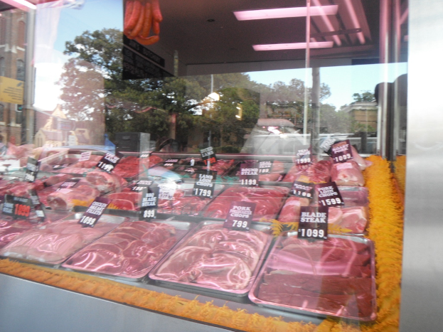 Eine Glasvitrine in einem Metzgerladen mit Tabletts voller Fleisch mit Preisschildern, mit Gebäuden und Bäumen durch das Fenster und der Decke des Raums oben im Bild.