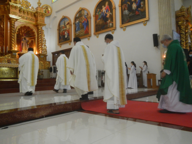 Eine Gruppe von Priestern geht einen roten Teppich in einer Kirche entlang, während Menschen auf dem Boden vor und rechts von ihnen stehen und Foto Rahmen an der Hintergrundwand zu sehen sind.