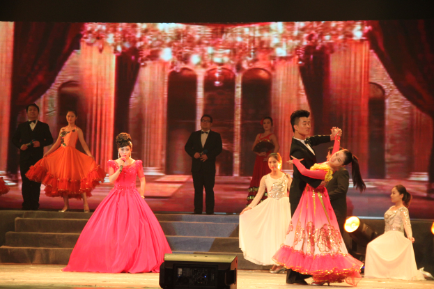 Eine Gruppe von Menschen steht auf einer Bühne mit Stufen und einem Hintergrundbild, beleuchtet von Scheinwerfern, mit einem Lautsprecher unten, was eine Tanzvorstellung vermuten lässt.