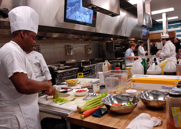 Eine Gruppe von Köchen in einer Küche, die Essen auf einem Schneidbrett zubereiten, mit einem Koch im Vordergrund in einer weißen Schürze und Mütze, der ein Messer hält und von Utensilien, Schüsseln, Gemüse, einem Handy, Flaschen und anderen Gegenständen umgeben ist, mit einem Fernseher und Deckenleuchten im Hintergrund.