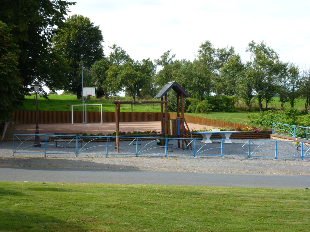Ein Park mit einem Spielplatz in der Mitte, umgeben von einer Straße, Gras, Pflanzen, Blumen, einem Zaun, Pfosten und einem Basketballkorb sowie Bäumen unter einem klaren blauen Himmel.