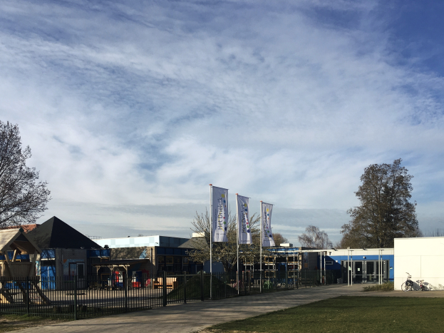 Ein großes Gebäude mit einem grasbewachsenen Bereich davor, umgeben von einem Metallzaun, einem Weg, Bäumen, Pfosten, Bannern, einem geparkten Fahrrad, nahen Häusern und einem bewölkten Himmel, das als neuer Spielplatz für Kinder an einer Schule dient.