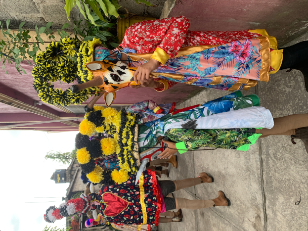 Eine Gruppe von Menschen in bunten Kostümen steht zusammen vor einem Gebäude, mit verschiedenen Gegenständen, Bäumen, Gebäuden und einem klaren blauen Himmel im Hintergrund bei einem Faschingsumzug.