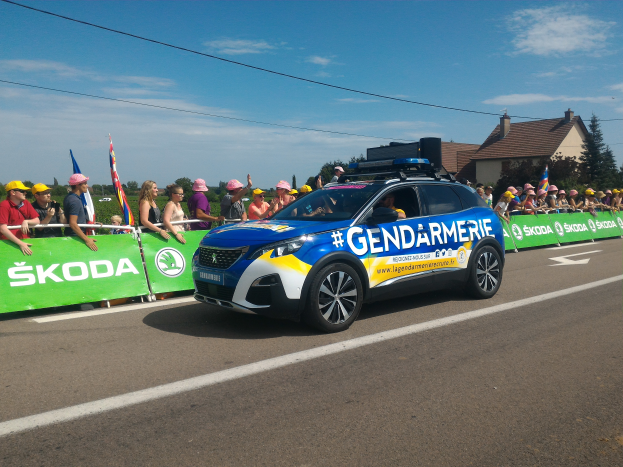 Ein Polizeiwagen fährt auf einer Straße neben einer Menge von Menschen, einige tragen Mützen und halten Fahnen, mit einem Banner auf dem Wagen und einer Kulisse aus Bäumen, Gebäuden und einem bewölkten Himmel. Der Text "Gendarmerie de la Tour de France" ist unten sichtbar.