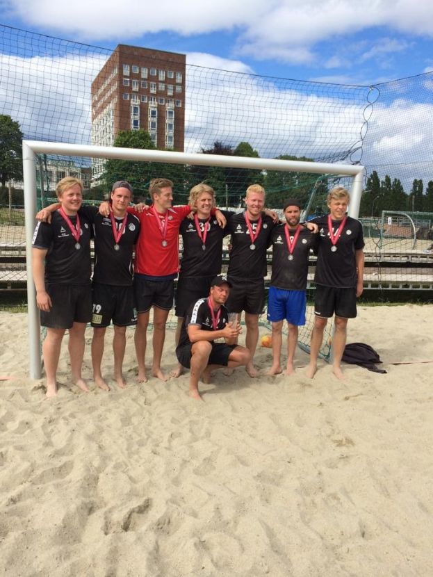 Gruppe von Männern in Sportkleidung auf einem sandigen Strand, mit Medaillen und umgeben von einem Netz, mit Bäumen, Gebäuden und einem bewölkten Himmel im Hintergrund.