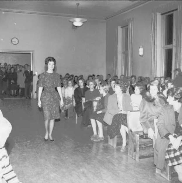 Eine Frau in einem schwarzen und weißen Kleid läuft über einen Laufsteg während einer Modenschau für Frauenrechte, mit einer Menge, die auf Stühlen sitzt und einer Uhr und Lampen an der Wand.