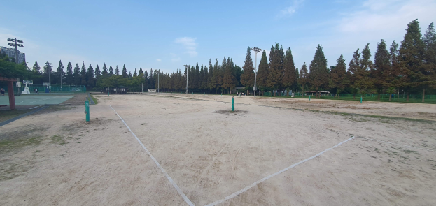 Ein Beachvolleyballfeld in einem Park, umgeben von Pfählen, einem Zaun, Bäumen, Gebäuden, Straßenlaternen und unter einem bewölkten Himmel.