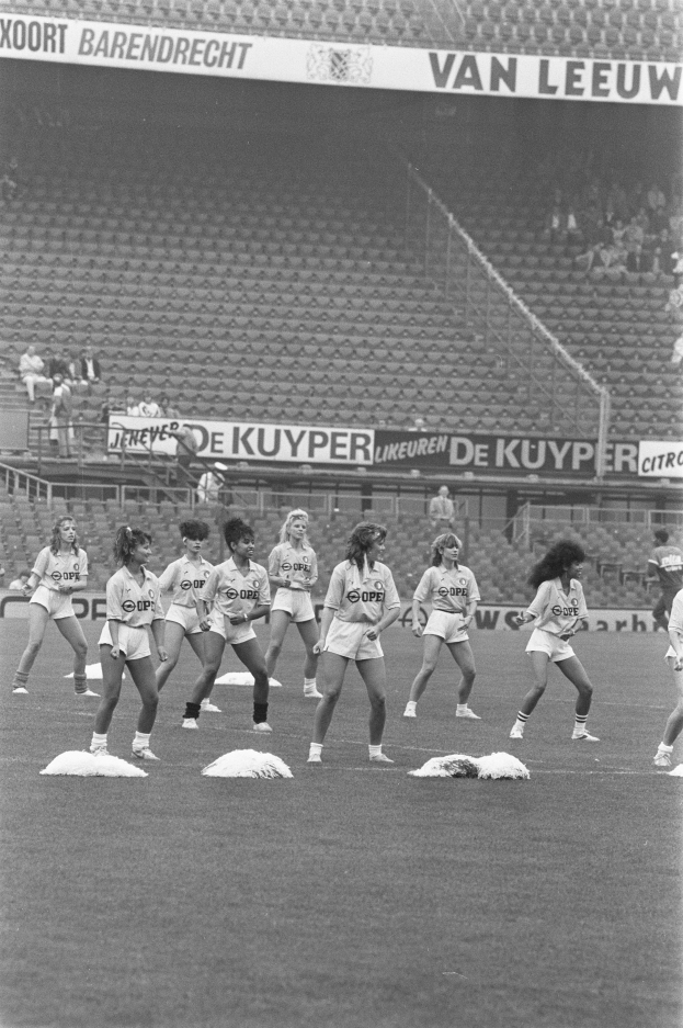 Eine Gruppe von Frauen auf einem Fussballfeld, umgeben von einer Menge im Publikum und Bannern mit Text, auf einem Schwarz-Weiß- Bild.