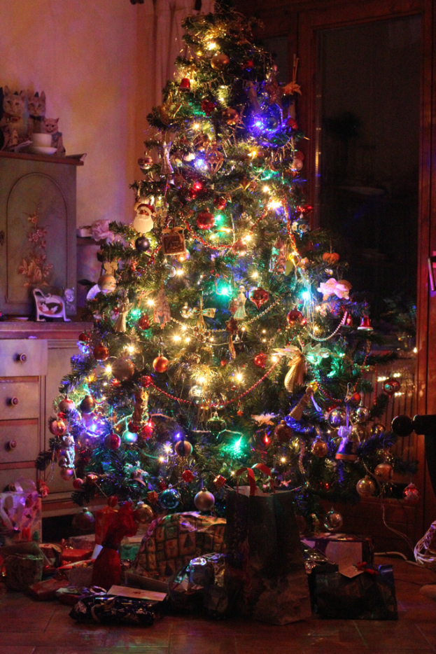 Ein Weihnachtsbaum mit Lichtern und Dekorationen, Geschenke auf dem Boden, ein Schrank mit Gegenständen auf der linken Seite und ein Fenster auf der rechten Wand.